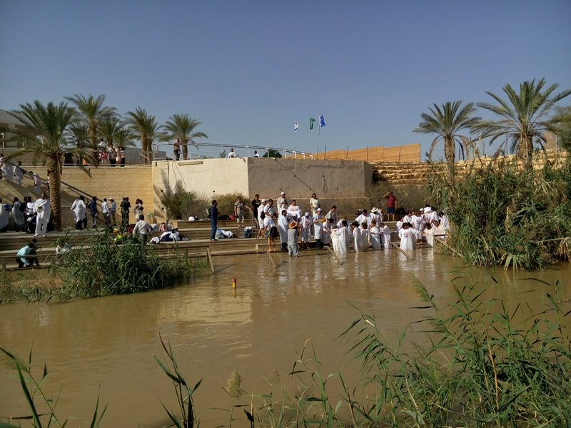 The Jordan River with palm trees and people gathered for a ceremony.