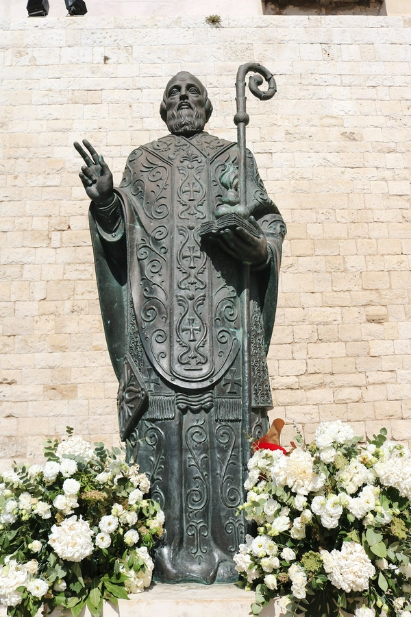 Statue of a religious figure at Basilica of Saint Nicholas, surrounded by colorful flowers and a stone wall.
