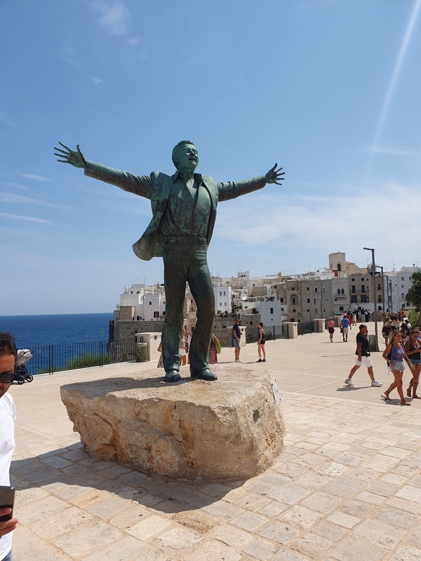 Statue of Domenico Modugno in Polignano a Mare with scenic cliffs and blue sea.